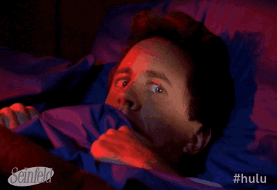GIF mostrar de maneira cômica homem deitado, segurando o cobertor até o queixo e olhando em volta com olhar assustado
